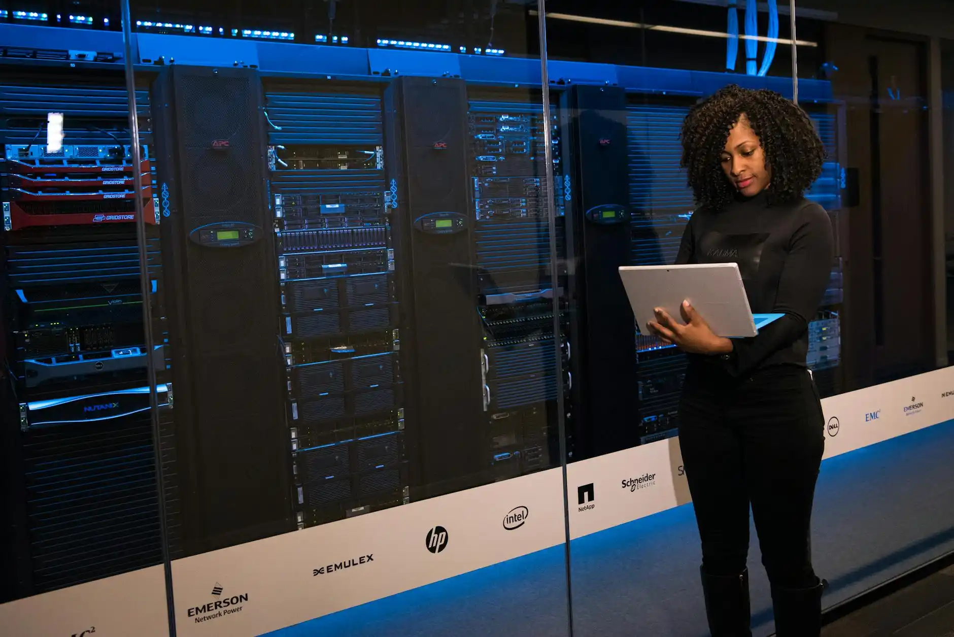 Engenheira em data center com racks de servidores e painéis de tecnologia, segurando um tablet em ambiente iluminado por luz azul, comparando sistemas legados e nuvem diante de tecnologia obsoleta.