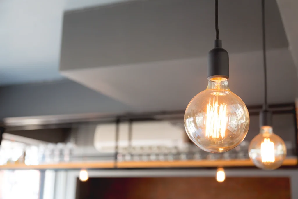 Lâmpada decorativa de filamento incandescente com globo de vidro acesa, pendente no teto, com outras luzes ao fundo. Ideal para ilustrar como escolher a iluminação ideal para cada ambiente da casa.