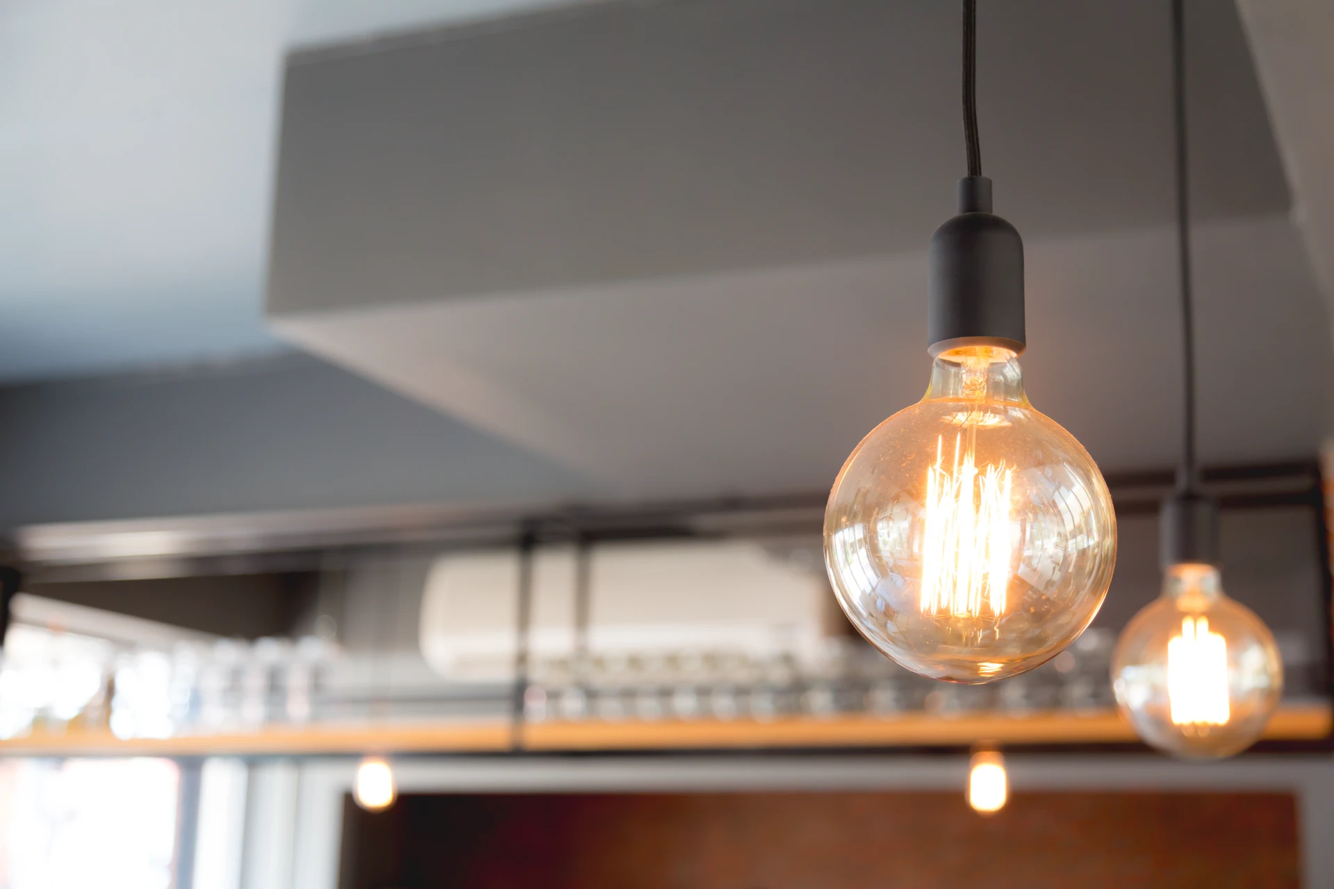 Lâmpada decorativa de filamento incandescente com globo de vidro acesa, pendente no teto, com outras luzes ao fundo. Ideal para ilustrar como escolher a iluminação ideal para cada ambiente da casa.