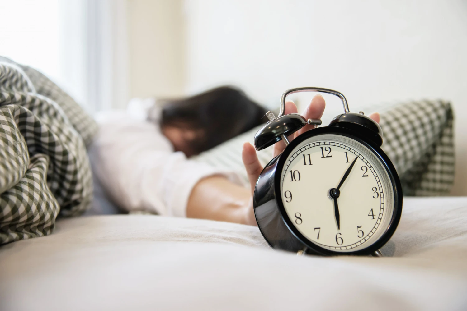 Relógio despertador preto sobre a cama de um quarto claro, com uma pessoa ao fundo de olhos fechados, sugerindo acordar com calma e reduzir o estresse diário usando minimalismo na prática.
