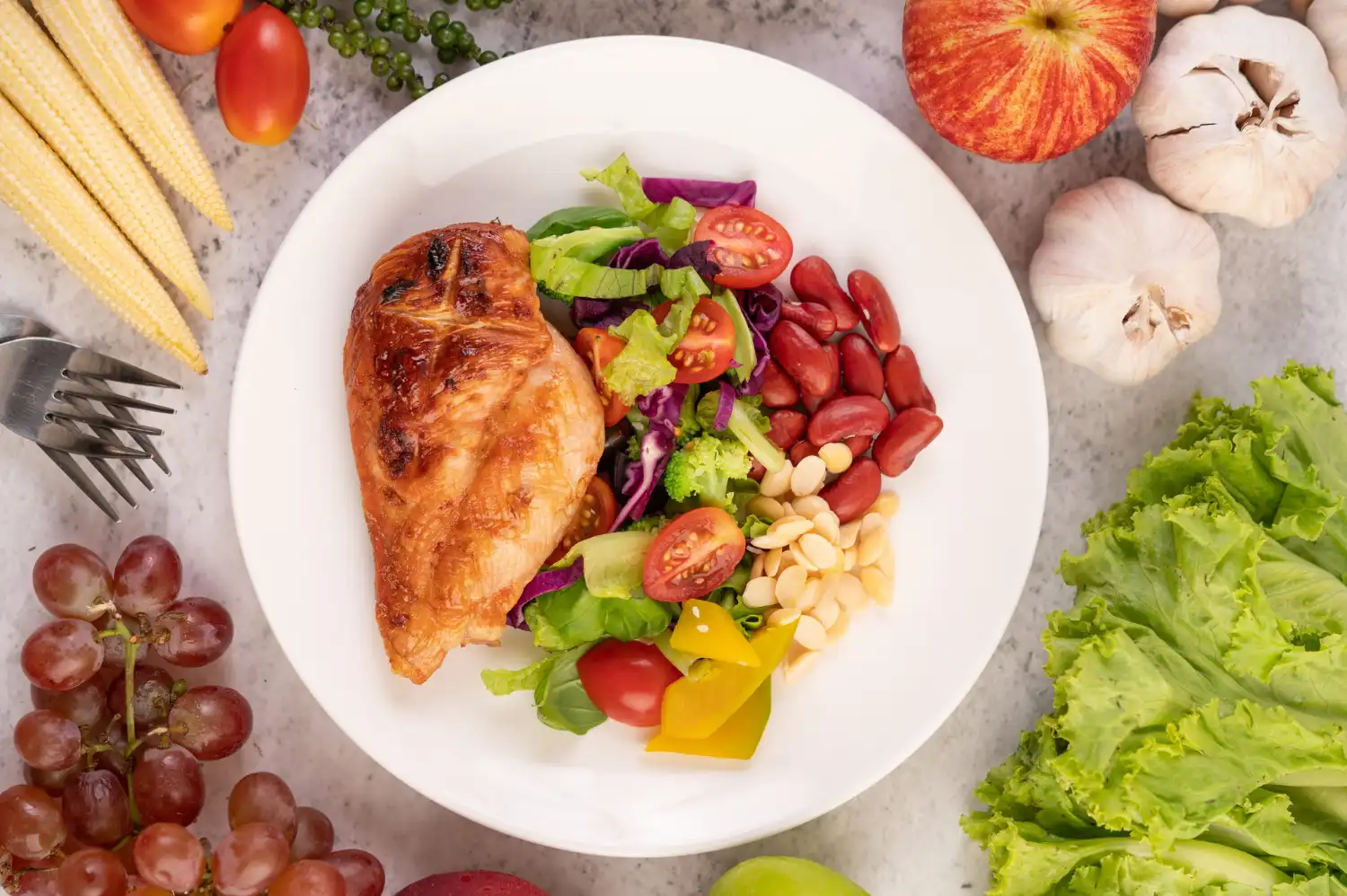 Prato com frango grelhado e salada variada com folhas verdes, tomate, pepino e uvas ao redor, em cenário culinário que remete à escolha de proteínas para ganho de massa muscular.