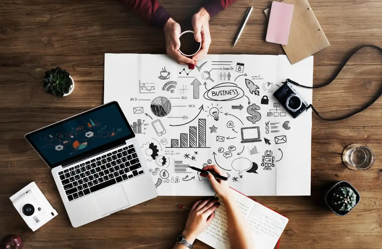 Pessoas analisam estratégias de negócio em papel com gráficos e ícones de marketing, enquanto trabalham em um laptop e câmeras sobre uma mesa de madeira; Marketing de Conteúdo: Como Criar Materiais que Atraem e Convertem Clientes.